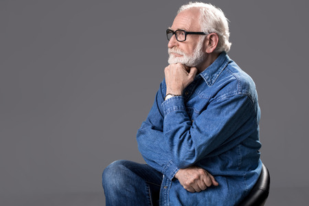 Profile of meditative male person in spectacles relaxing on stool. He is looking aside with calm face. Copy space in left side. Isolated on grey backgroundの写真素材