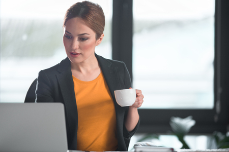 Portrait of happy young girl working with notebook computer while tasting mug of delicious coffee in apartment. Occupation concept. Copy spaceの写真素材