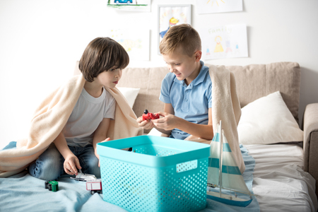 Two joyful young boys are sitting on sofa with box full of toys and looking on plastic figure, one of them is holding in hand with excitementの写真素材