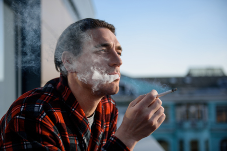 Calm adult man puffing on a cigarette with thoughtful look, he is staying on terrace. Focus on his faceの写真素材