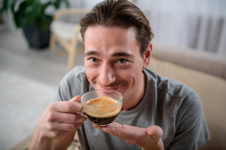 This is delicious. Portrait of calm male enjoying coffee and smiling. He is holding a cup in his hands and looking at camera with happinessの写真素材