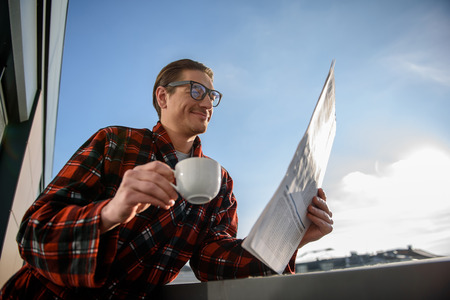Low angle of satisfied adult male reading daily newspaper with glad smile, holding a cup in his hand. He is standing on the balconyの写真素材