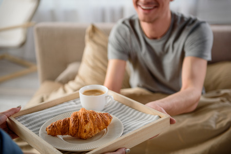 Close up of male and female arms holding tray, focus on salver with coffee and croissant. Man is smiling and sitting in bedの写真素材
