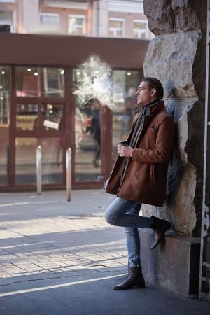 Full length side view pensive male drinking cup of delicious liquid while leaning against wall on street and smoking. Dreaminess conceptの写真素材