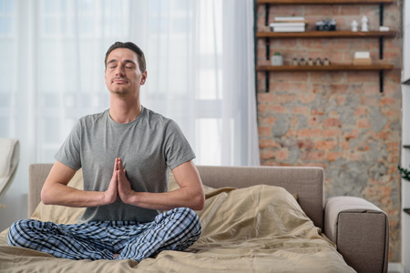 Feeling calm. Serene adult man keeping eyes closed  and sitting on bed in a lotus position. Copy space in right sideの写真素材