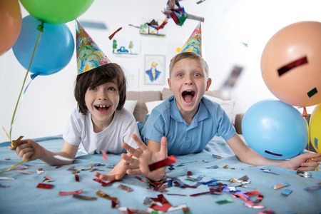 Portrait of two jolly youngsters in living room holding colorful balloons, children are surrounded...