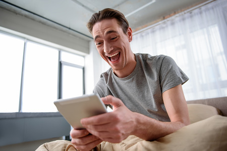 Portrait of happy adult male sitting on bed and looking at tablet with smile. Low angleの写真素材