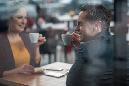 Side view outgoing man tasting cups of liquid while locating at desk in cafeteria. He telling with happy female. Coffee break conceptの写真素材