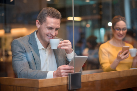 Portrait of cheerful male watching at electronic tablet. Happy girl typing in mobile. They tasting mugs of tea while situating at desk. Relax conceptの写真素材