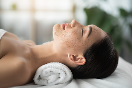 Calm young woman is relaxing at beauty salon. She has peeling mask on her face. Her eyes are closed with pleasureの写真素材
