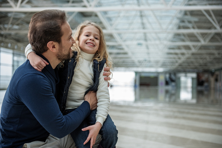 Pleased man and child standing at the airport. They are in embrace while girl is sitting on his lap. Copy space in right sideの写真素材