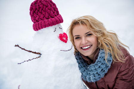 Portrait of cheerful young woman embracing snowman with joy and laughing. She is looking at camera with happiness. Winter entertainment conceptの写真素材