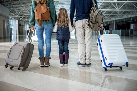 Low angle of adults standing at the airport after disembarkation. They are keeping few suitcases and their kid close to themselves. Focus on their backの写真素材