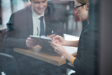 Smiling businessman and calm female watching at appointment book. They sitting at desk indoor. Career and discussion conceptの写真素材