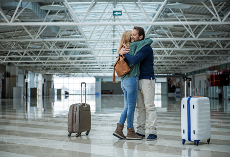 Two cheerful adults hugging each other before departure while standing in the terminal. Suitcases are apart of themの写真素材