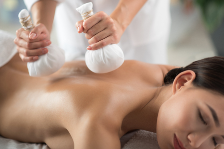 Close up of masseuse hands massaging female back by herbal pouchesの写真素材