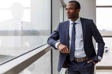 Feeling relaxed. Elegant experienced adult african businessman in suit is standing and looking through the window thoughtfully while holding hand in pocket of his pants. Copy space in the left sideの写真素材