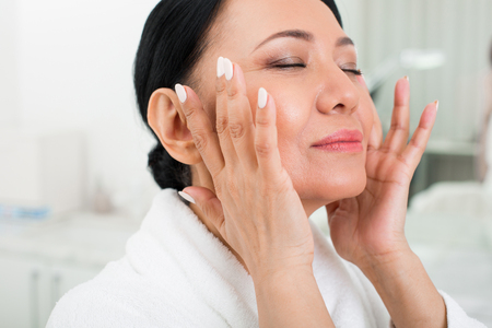 Feeling good. Calm senior woman using cosmetic at spa. She is rubbing the cream with her finger. Her eyes are closedの写真素材