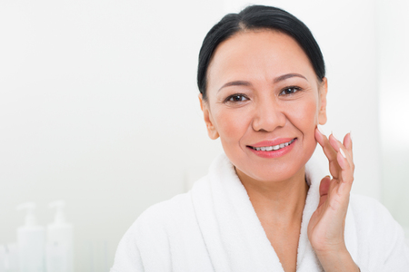 Portrait of middle-aged asian woman looking at camera. She is smiling and feeling her skin at spa. Copy space in left sideの写真素材