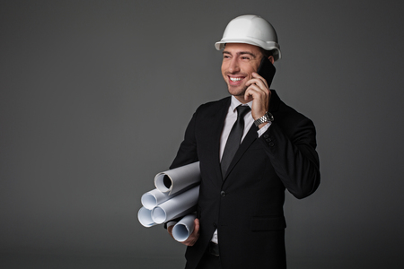 Portrait of cheerful unshaven builder telling by mobile while keeping different projects in hand. Communication and work concept. Isolatedの写真素材