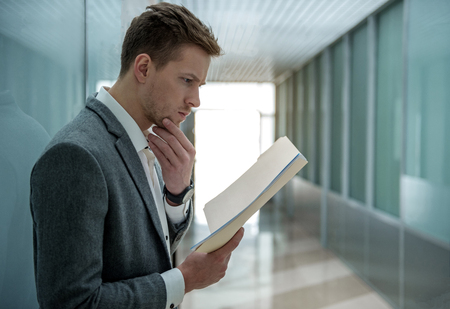 Busy morning. Pensive young professional employee is reading documents with concentration. He is standing in hallway and touching his chin. Copy space in the right sideの写真素材