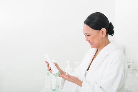 Pleased asian female looking at tube of cream in her hand. She is examining the instruction in beauty salon. Copy space in left sideの写真素材