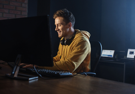 Side view happy unshaven programmer writing on keyboard of computer while sitting at table....