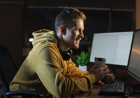 Side view cheerful bearded man drinking cup of coffee while typing in keypad of modern digital...