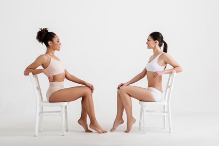 Side view profile of satisfied women being proud of their slimness. They are resting in underwear facing each other. Isolated on backgroundの写真素材