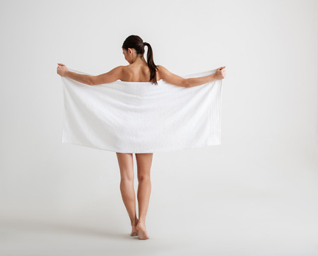 Undressed calm girl standing with her back. She is hiding her graceful body behind a towel. Isolated on backgroundの写真素材