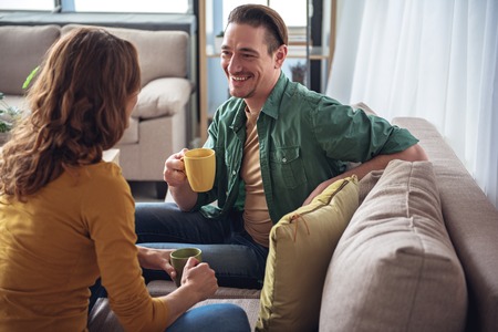Cheerful married couple resting on sofa in living room together. They are drinking tea and laughing. Pleasant conversation conceptの写真素材