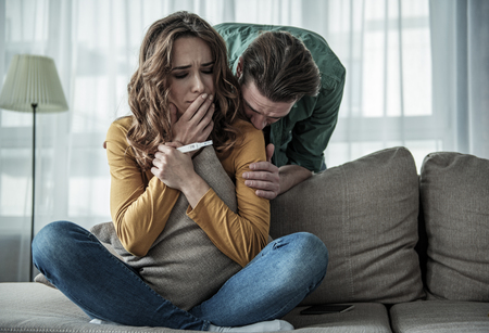 I am pregnant. Portrait of worried young woman holding pregnancy test stick while sitting on couch. Man is kissing her with loveの写真素材