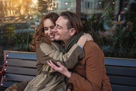 Happy loving couple is hugging with fondness. They are sitting on bench and laughingの写真素材