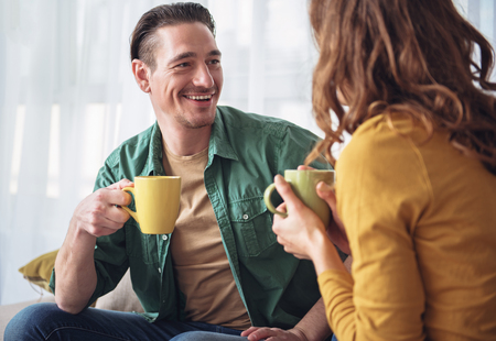 Portrait of excited young man talking with his wife at home. He is drinking tea and smiling. Couple is sitting on couchの写真素材
