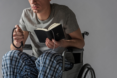 Concentrated disabled guy taking seat in armchair with holy book and beads in arms. Isolated on backgroundの写真素材