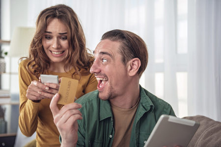 Excited man is showing cold credit card to his wife and laughing. Woman is typing on smartphone quickly while ordering things in internet. Online shopping conceptの写真素材