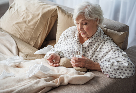 Upset old woman sitting in bed with sad look. She is looking at medication in hands and holding glass of water. Copy space in left sideの写真素材