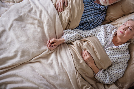Top view of tranquil pensioners sleeping in bedroom. They are holding hands of each otherの写真素材