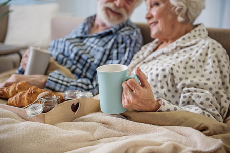 Focus on the female hand holding cup of coffee. Glad old woman and man relaxing in bedroom. They are eating croissantの写真素材