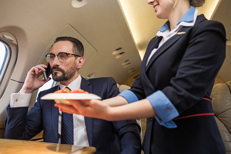 Portrait of thoughtful good looking mature man talking by phone in airplane. Stewardess is serving...