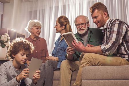 Happy woman taking with grandmother on couch opposite boy typing in electronic tablet. Smiling grandfather and male looking at frame. Entertainment conceptの写真素材