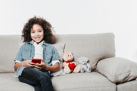 Portrait of cheerful little girl relaxing on sofa with lovely toys and smartphone in hand. She is looking at camera with joy. Copy space on right side. Isolated on backgroundの写真素材