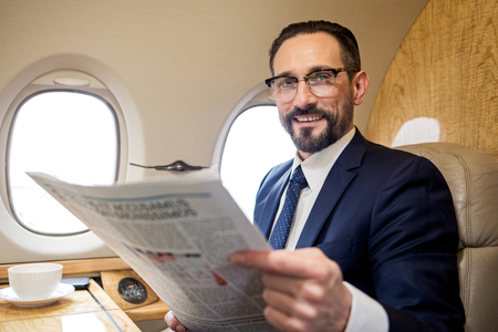 Portrait of pleased bearded male sitting in airplane seat and holding paper, he is looking at camera...