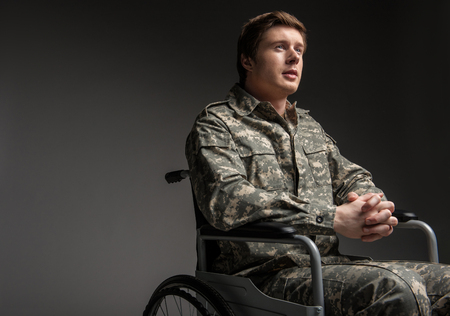 Cheerful disabled young military man looking up with hope. He is sitting in wheelchair with his hands folded in the lock. Isolated on grey background. Copy space in left sideの写真素材