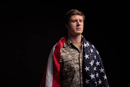 Waist up portrait of serious male soldier standing covered in usa flag. He is looking with sadness. Isolated on background. Copy space in left sideの写真素材