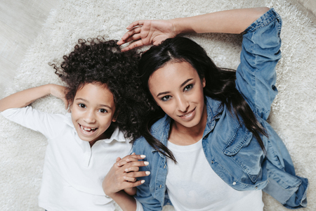 Top view portrait of smiling mom and daughter reposing on soft carpet and holding hands. They are looking at cameraの写真素材