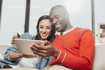 Waist up portrait of content man and woman resting at home in embrace and using tab togetherの写真素材