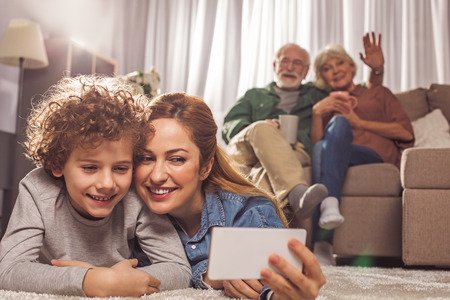Portrait of happy female and smiling kid taking selfie by mobile while locating on carpet. Beaming grandparents drinking cup of tea and sitting on sofa. Entertainment conceptの写真素材