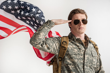 Portrait of sedate male soldier returning a salute with his right hand. American Flag is fluttering behind his back. Concept of pride. Isolated on backgroundの写真素材