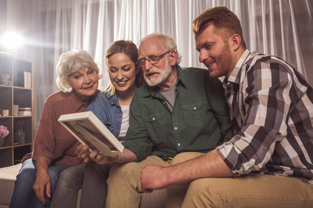 Happy granny with beaming lady and outgoing grandfather with man looking at frame in apartment. Memory conceptの写真素材